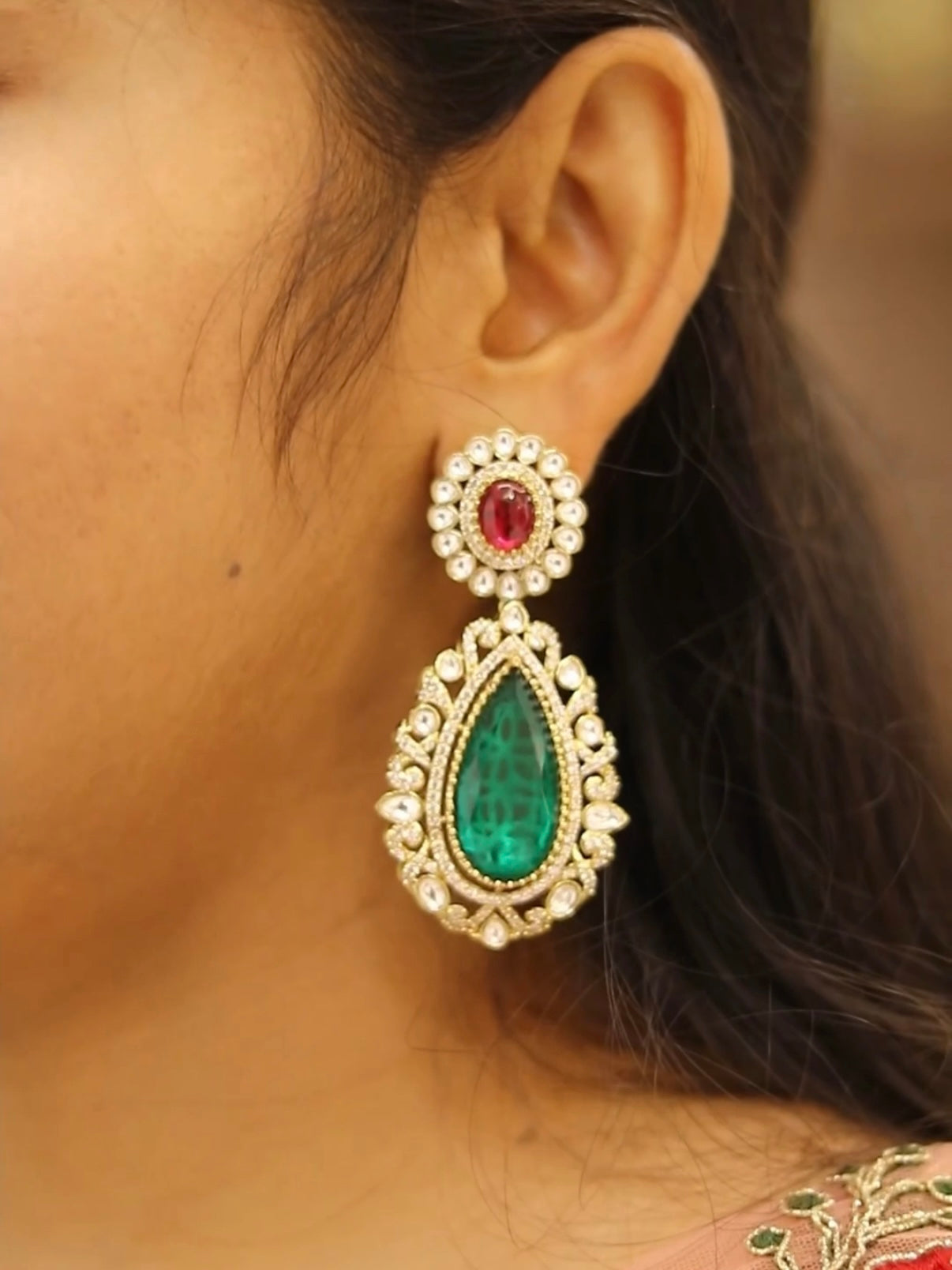 Close-up of a person wearing a decorative earring with green gemstone and red stone.
